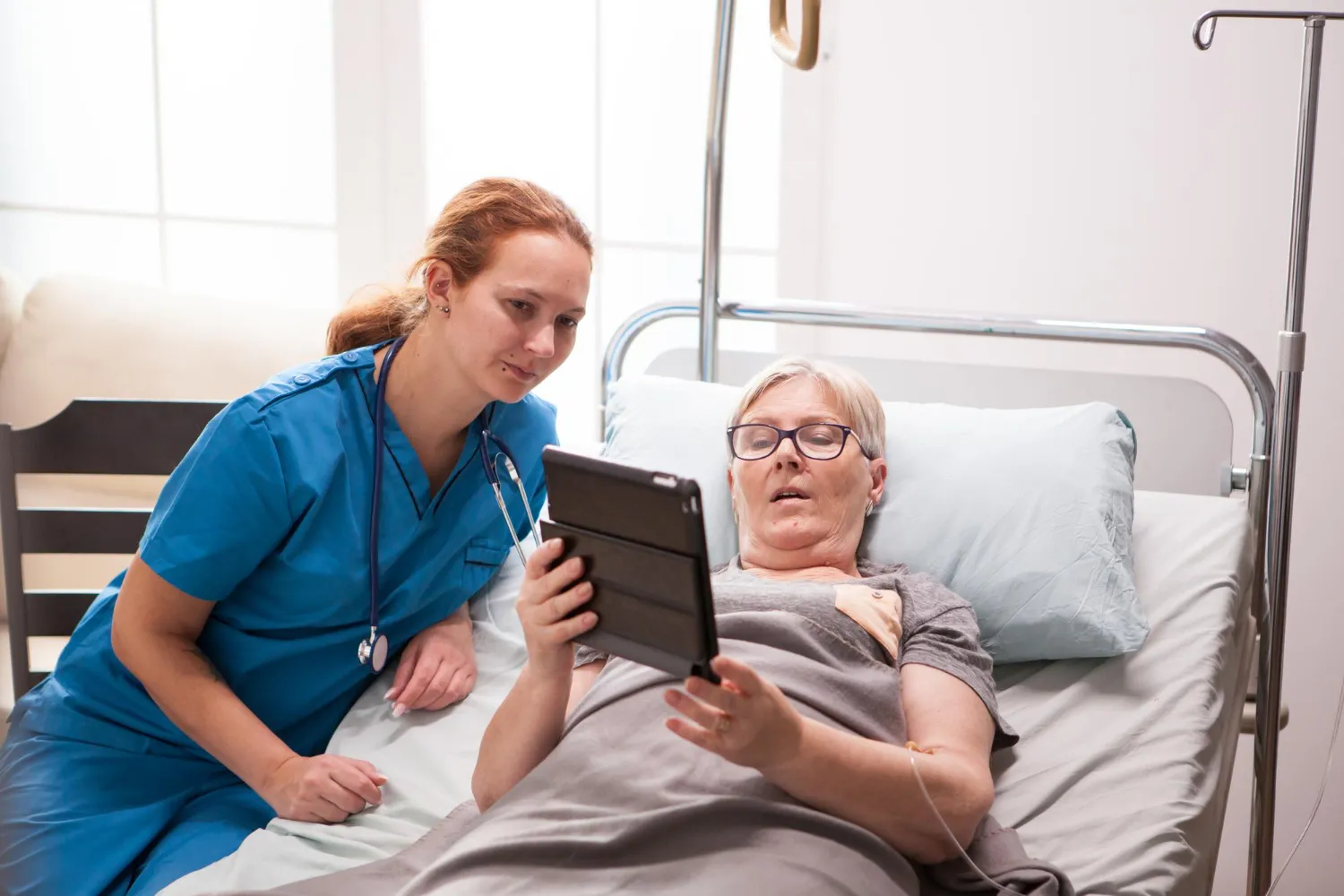 Nurse assisting an elderly patient using a tablet in a hospital bed, representing transitional care management and the use of digital tools for effective patient care management after discharge