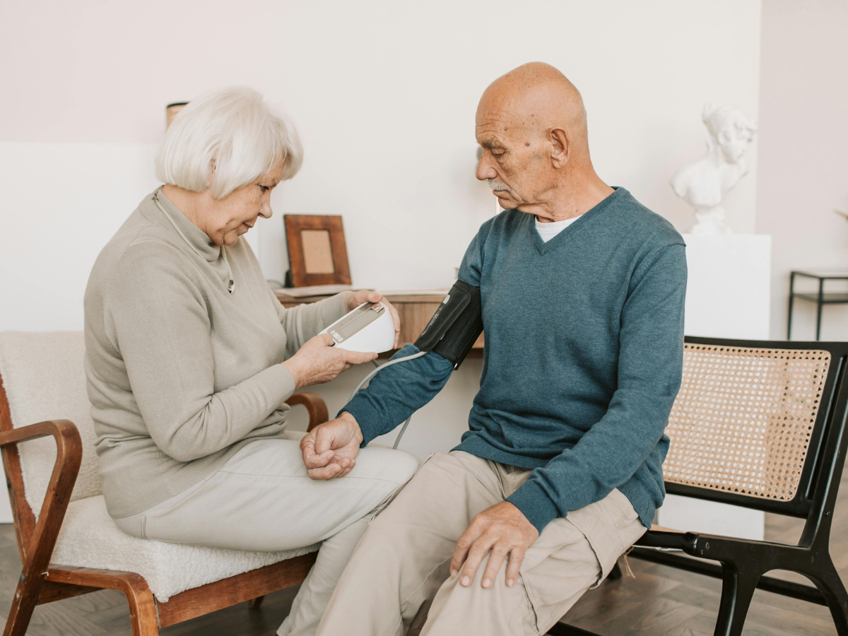 An elderly woman measuring the blood pressure of an elderly man at home using a digital blood pressure monitor, showing routine health monitoring, senior care, and supportive at-home medical assistance in a comfortable living environment