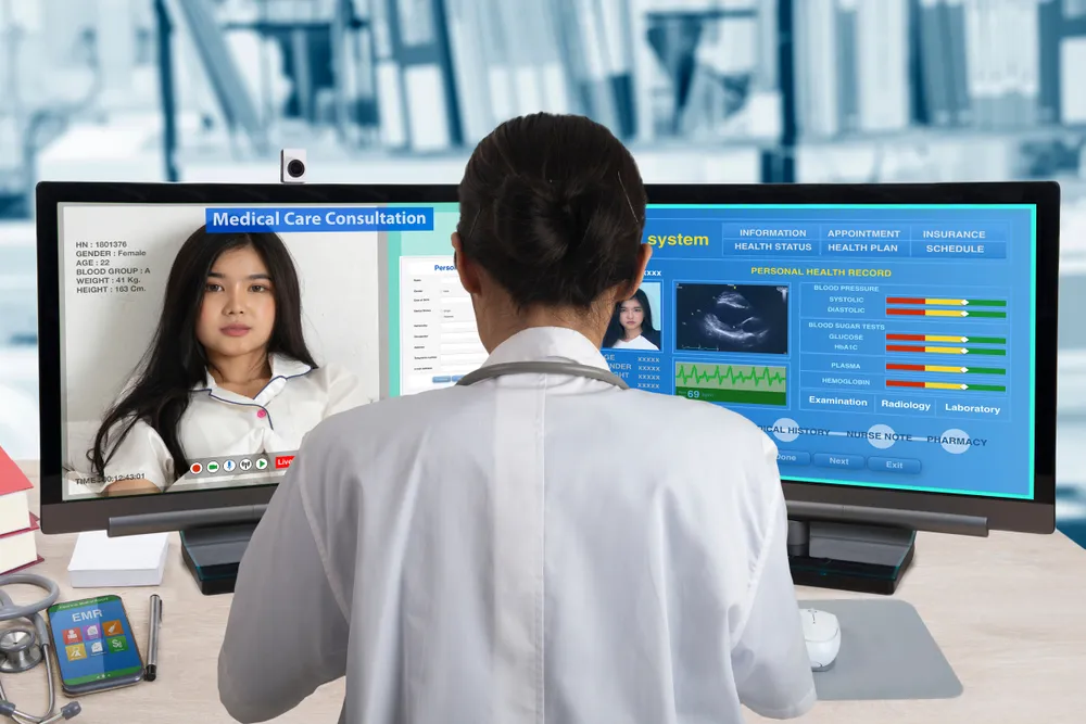 Doctor conducting a telehealth consultation on dual monitors showing a patient video call and electronic health records with charts and vitals, representing modern remote healthcare, digital diagnostics, and connected medical services.