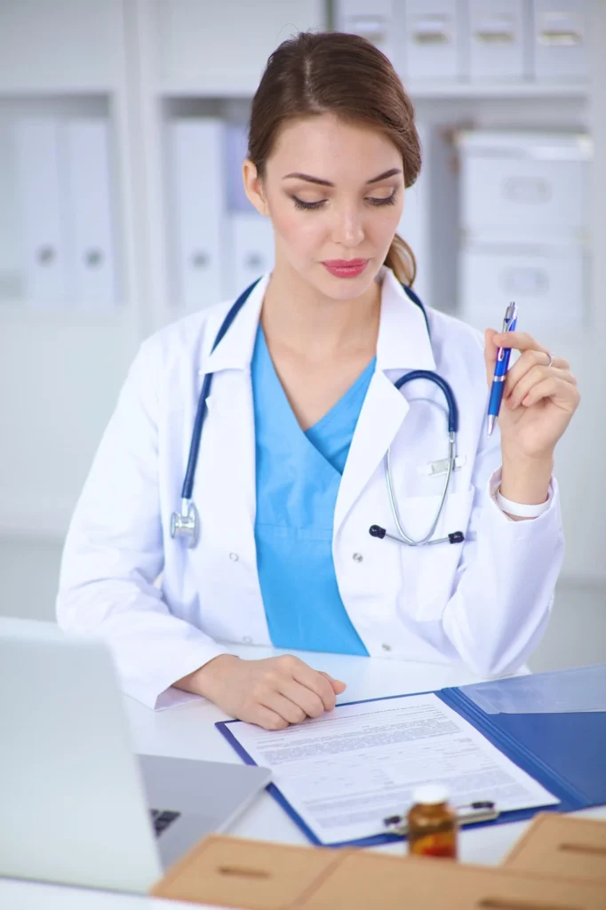 A female doctor in a white coat and blue scrubs with a stethoscope around her neck sits at a desk, reviewing and writing on a document on a clipboard while holding a pen. A laptop, medication bottle, and binders are visible nearby, suggesting a clinical documentation or prescription setting.