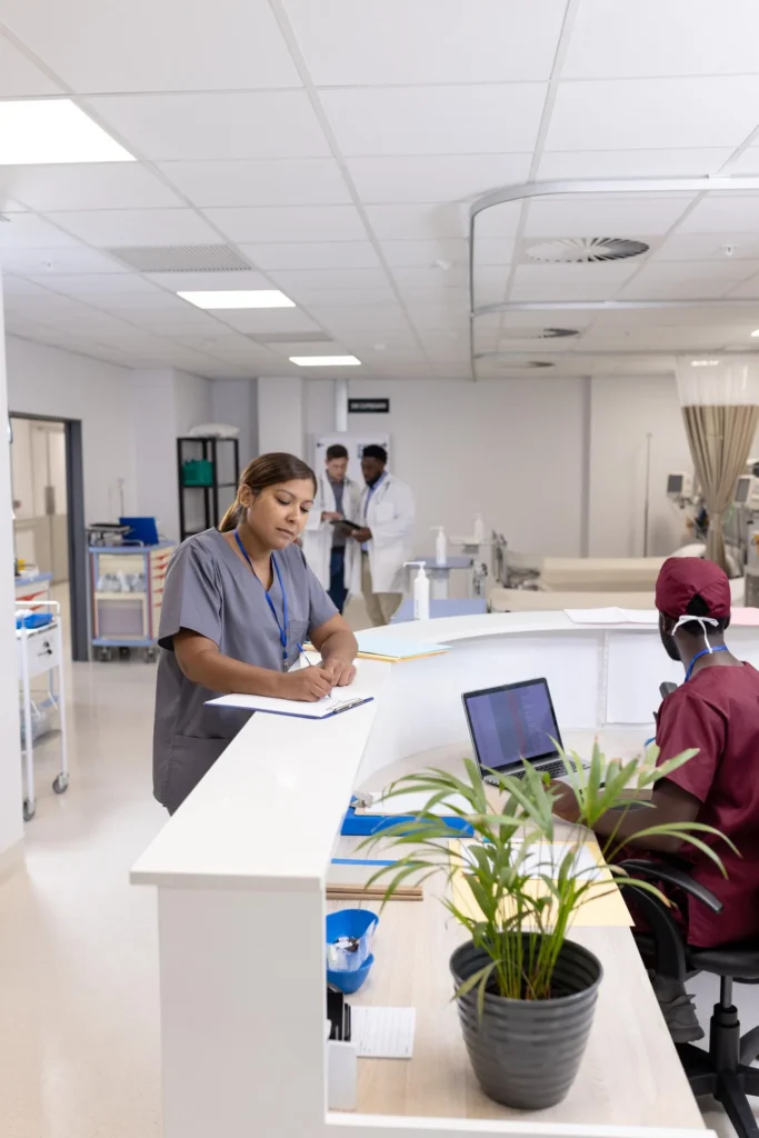 Busy hospital nurse station with healthcare staff working on a clipboard and laptop, while doctors consult in the background, representing coordinated patient care, clinical teamwork, and a modern medical environment.