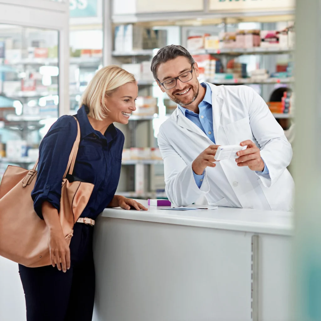A male pharmacist in a white coat and glasses smiles while showing a medication package to a female customer with a handbag at a pharmacy counter, with shelves of medications visible in the background, representing a friendly pharmacist-patient consultation.