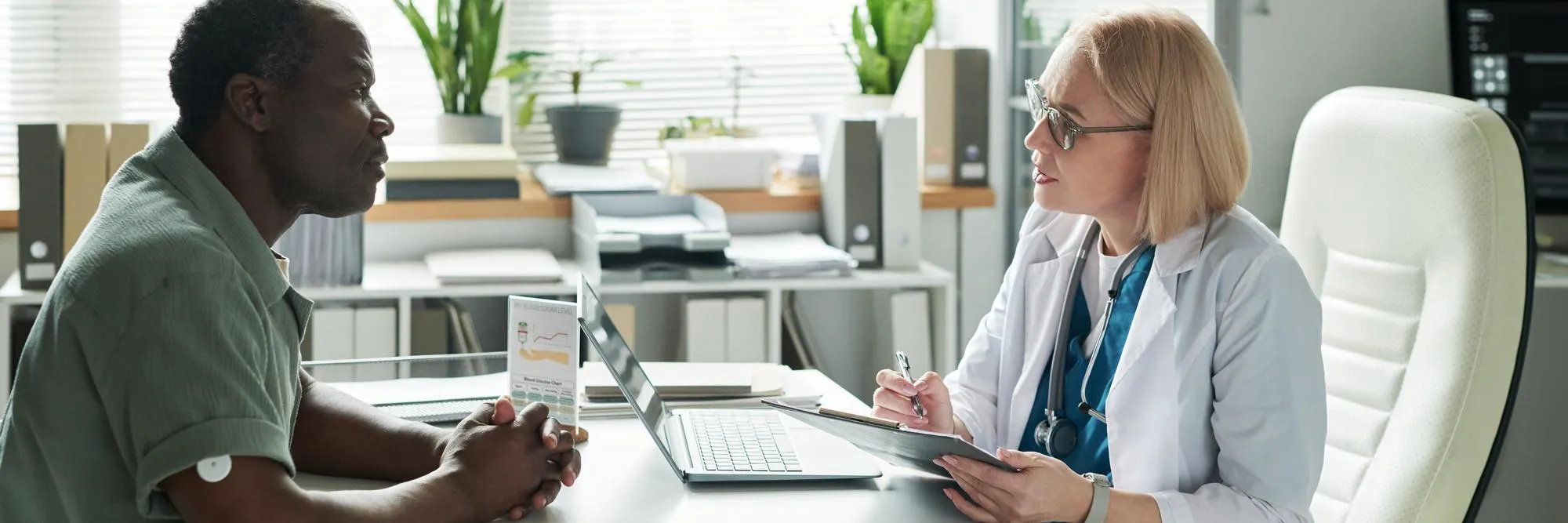 Doctor in a white coat consults with a patient at a desk, with a laptop and charts nearby on a bright office.