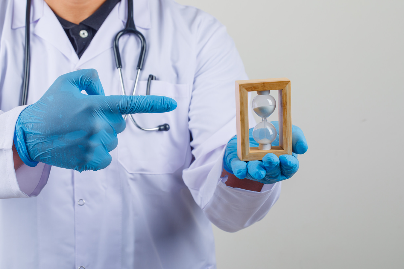 Medical professional in a white coat with blue gloves points to a wooden-framed hourglass he is holding, signaling time.